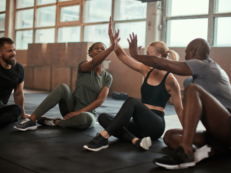 Four diverse individuals in a warmly lit private gym who are reflecting on their unique body transformation journeys, highlighting hope, individuality, and authentic progress after a workout, with visible sweat, textured activewear.